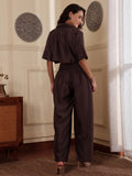 Back shot of a Woman wearing a brown co-ord set with crop shirt and flared Pant outfit standing in a room with a neutral wall and window.