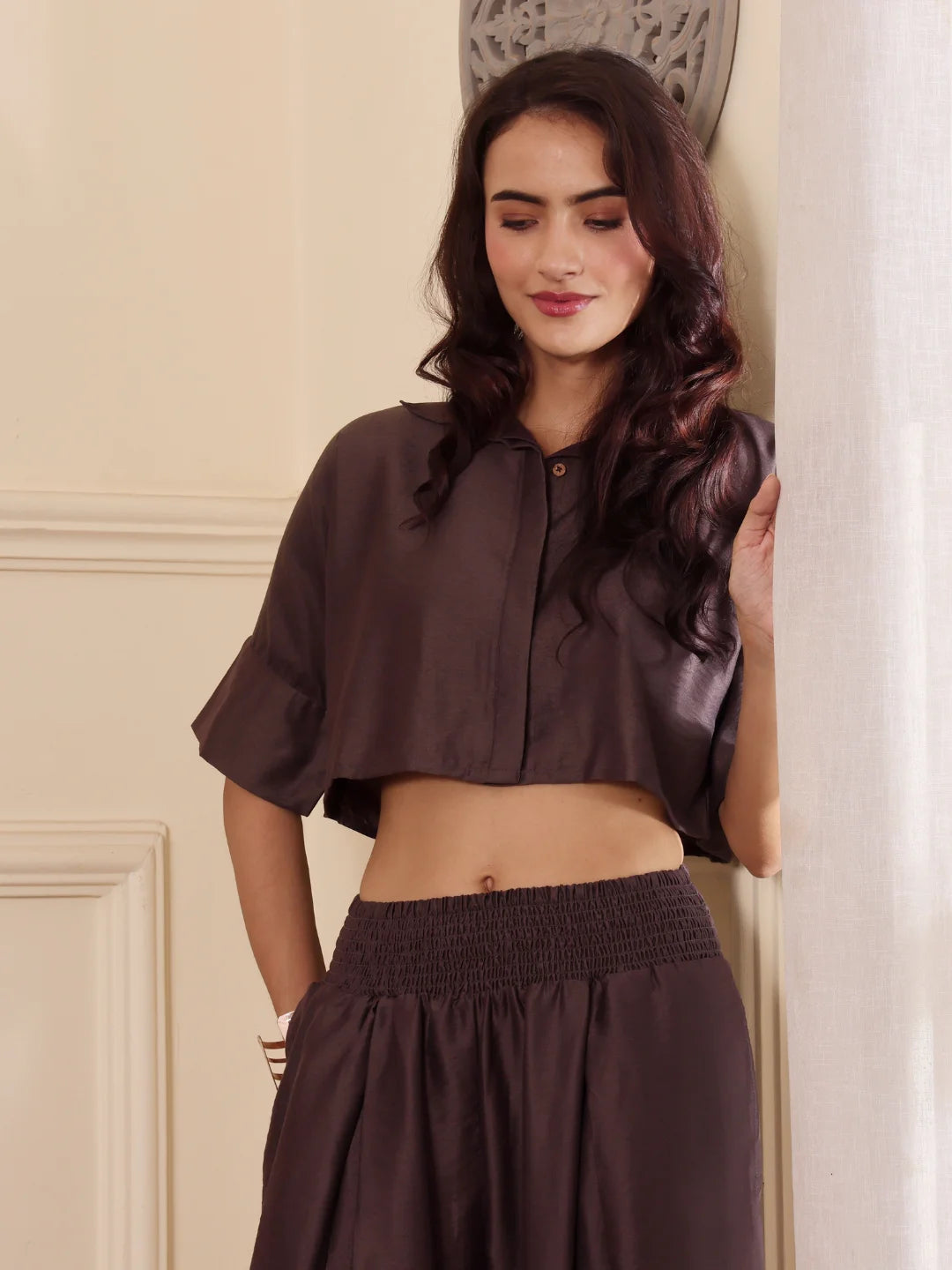 Close up shot of a woman wearing a brown co-ord set with crop shirt and flared Pant outfit standing in a room with a neutral wall and window.