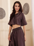 Front shot of a Woman wearing a brown co-ord set with crop shirt and flared Pant outfit standing in a room with a neutral wall and window.