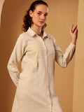 A woman modeling a light beige linen shirt dress with tonal floral embroidery on the front, posed against a warm beige background.