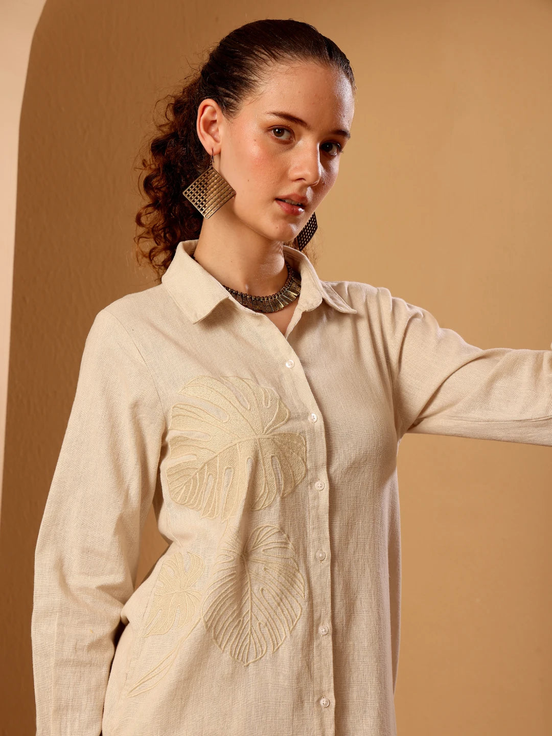 A woman wearing a light beige linen button-up shirt with tonal embroidered leaf patterns on the front, posed against a warm beige background.