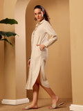 A woman models a long-sleeved, button-up linen midi dress in a warm cream color, standing in a warm beige architectural setting with arches and a potted plant.