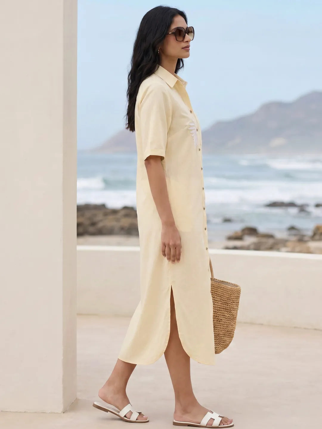 Side profile of a Woman in a cotton linen blend yellow shirt dress with embroidery holding a straw bag by a beach.