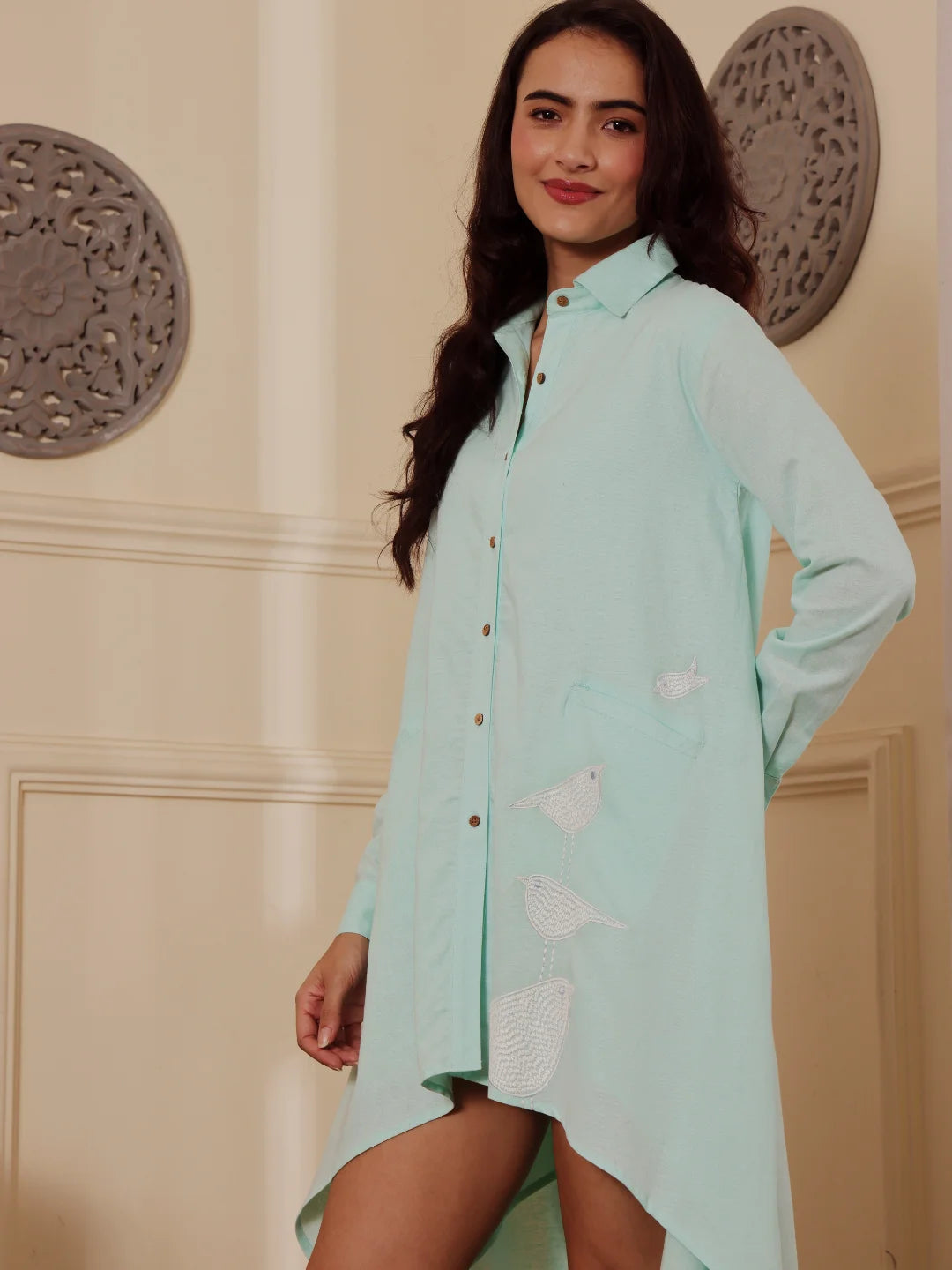 Side profile of a woman wearing a mint blue linen blend Shirt dress with embroidery and a patterned wall in the background