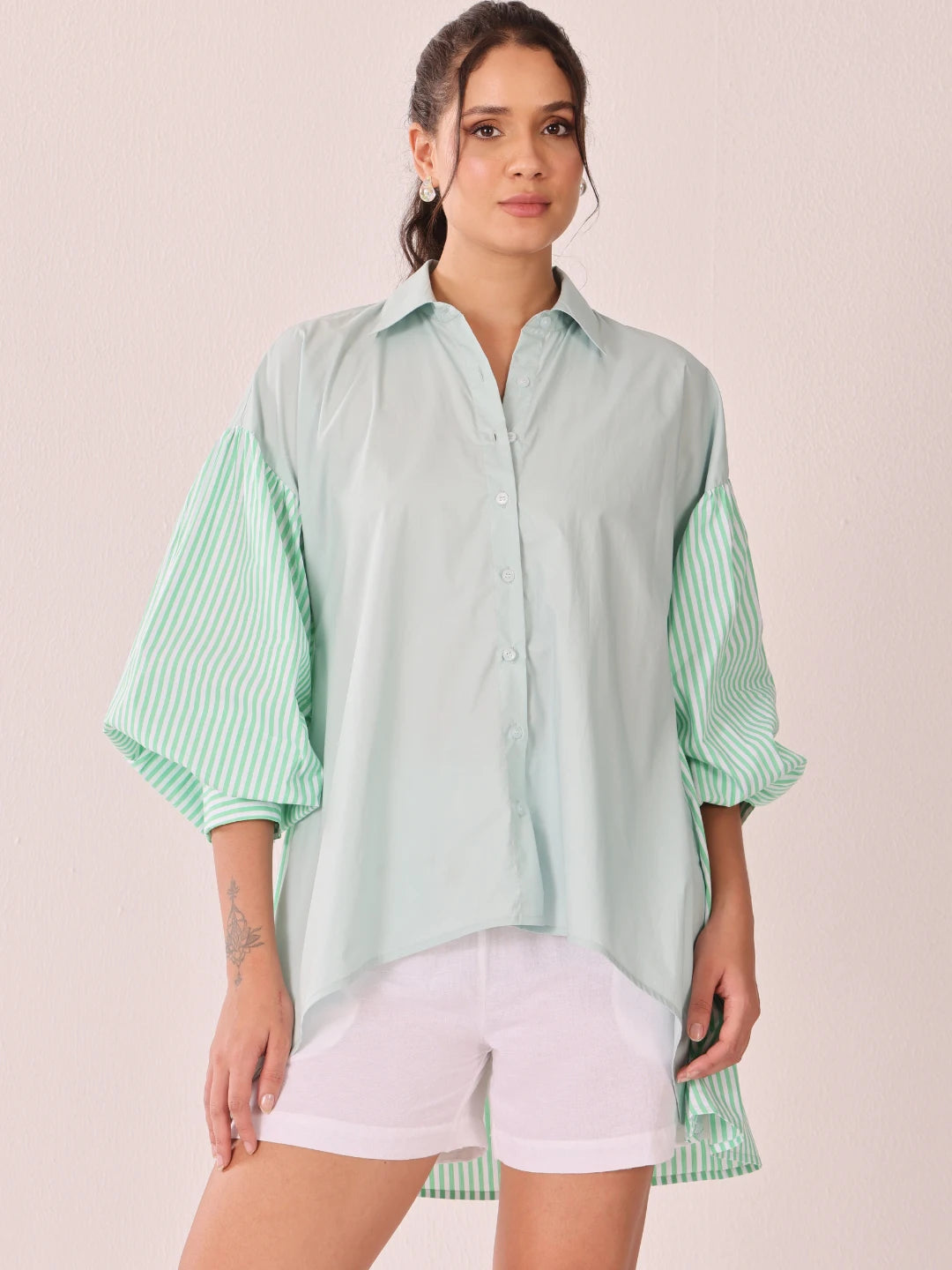 A woman modeling a mint green oversized button-up shirt with contrasting striped balloon sleeves, paired with white shorts, against a solid pale pink background.