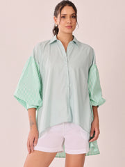 A woman modeling a mint green oversized button-up shirt with contrasting striped balloon sleeves, paired with white shorts, against a solid pale pink background.