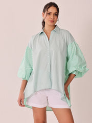 A woman modeling an oversized button-up shirt with a pale mint green body and green-and-white vertically striped balloon sleeves, paired with white shorts against a solid pale pink background.