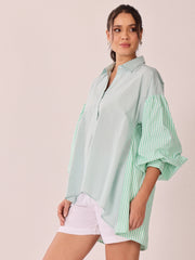 A woman modeling an oversized button-up shirt with a pale mint green body and green-and-white striped bell sleeves, paired with white shorts, against a plain light beige studio background.