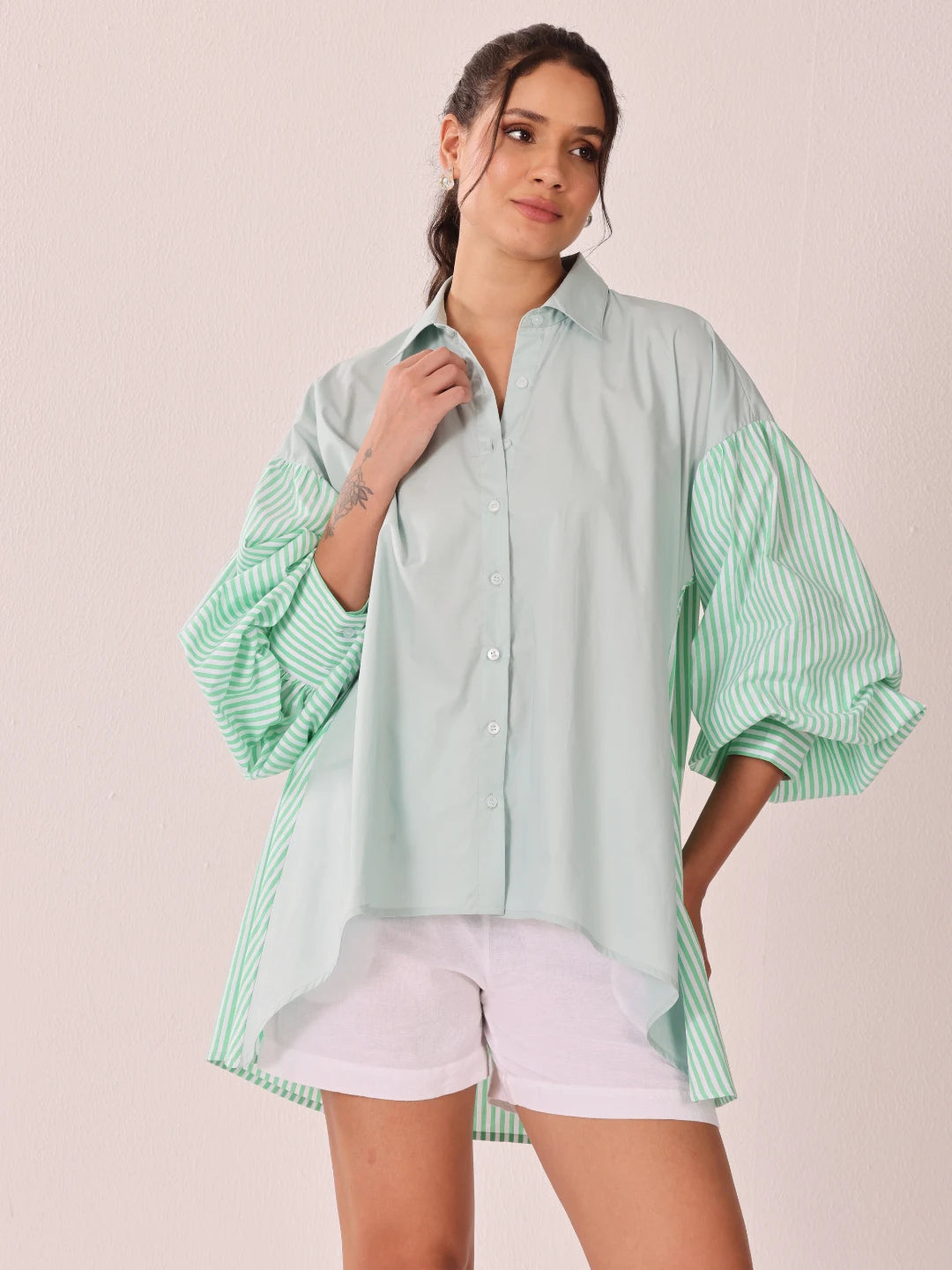 A woman modeling a light mint green button-up shirt with voluminous striped balloon sleeves and white shorts against a solid pale pink background.