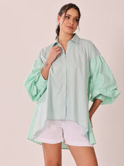 A woman modeling a light mint green button-up shirt with voluminous striped balloon sleeves and white shorts against a solid pale pink background.