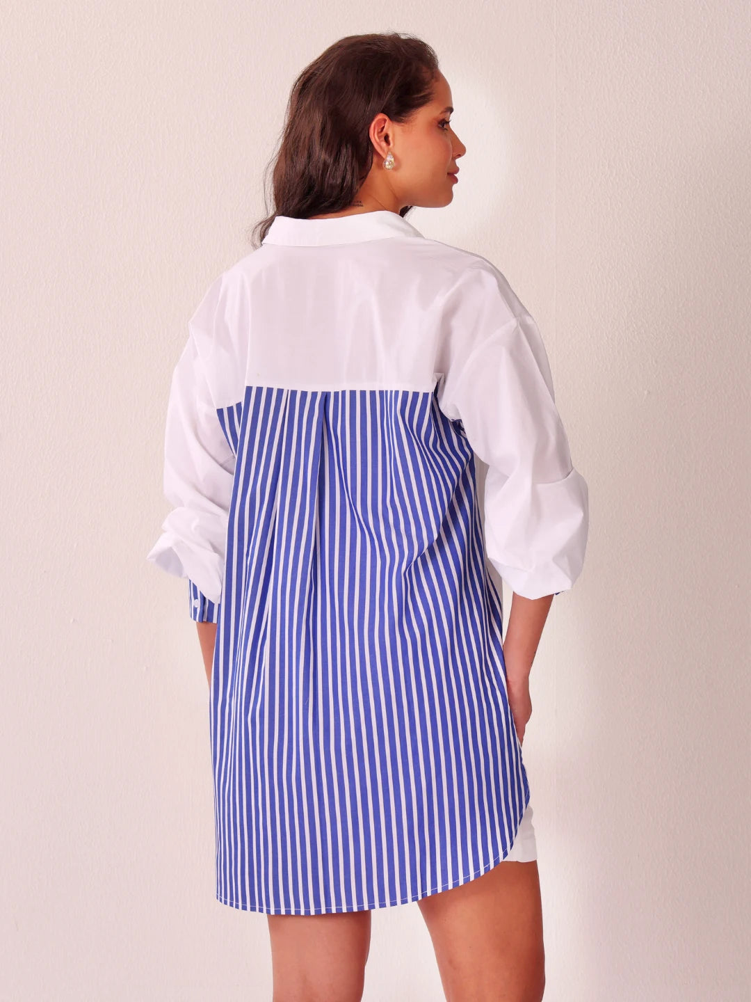 A woman shown from the back wearing a color-block shirt dress with a solid white upper back and collar, and a blue and white vertically striped lower back panel, against a plain off-white background.