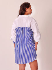 A woman shown from the back wearing a color-block shirt dress with a solid white upper back and collar, and a blue and white vertically striped lower back panel, against a plain off-white background.