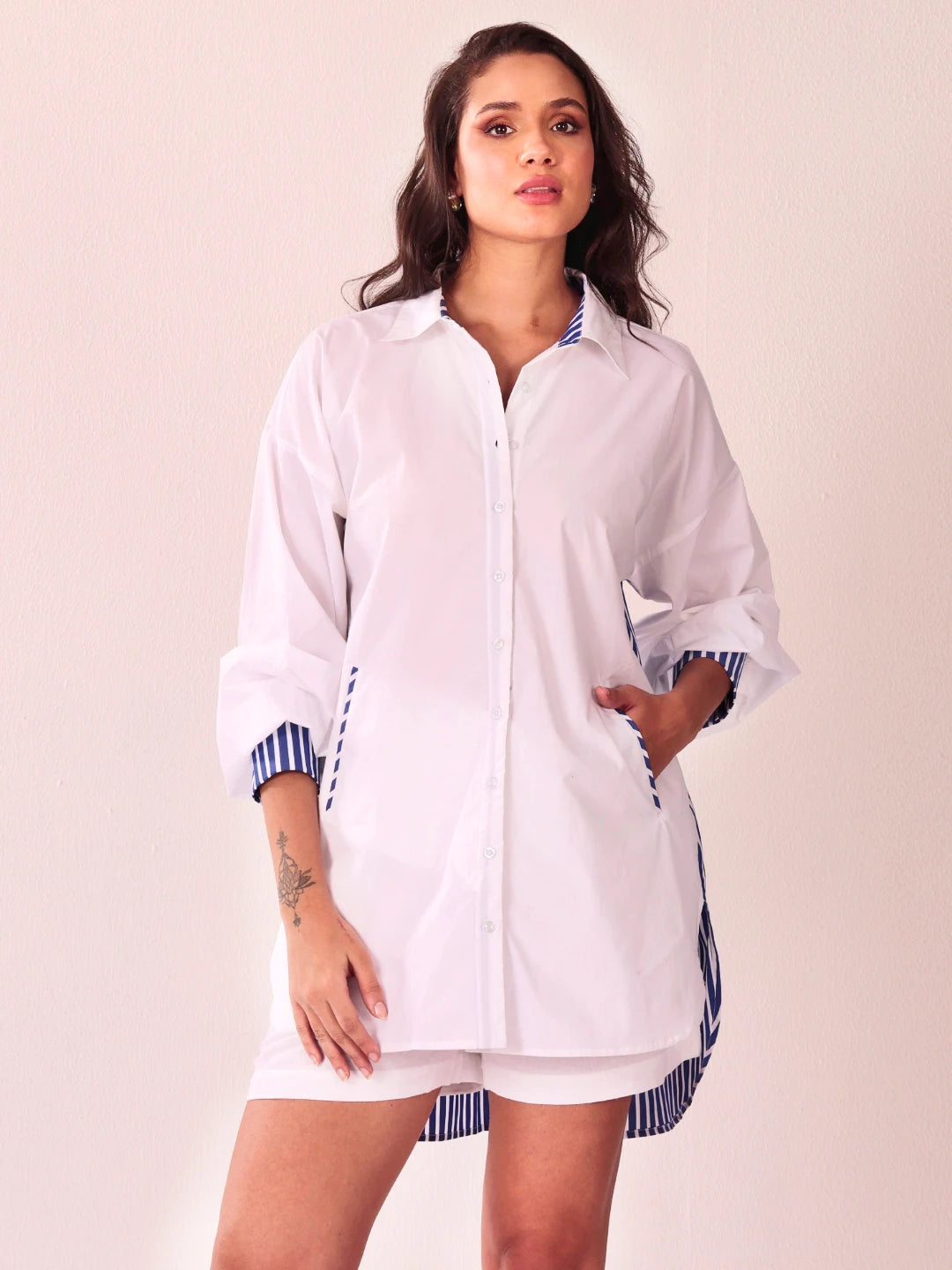 A woman modeling a white button-up shirt dress with blue and white striped trim on the collar, cuffs, and hem, paired with matching white shorts, against a solid light beige background.