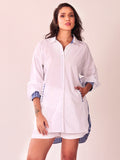 A woman modeling a white button-up shirt dress with blue and white striped trim on the collar, cuffs, and hem, paired with matching white shorts, against a solid light beige background.