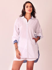 A woman modeling a white button-up shirt dress with blue and white striped trim on the collar, cuffs, and hem, paired with matching white shorts, against a solid light beige background.