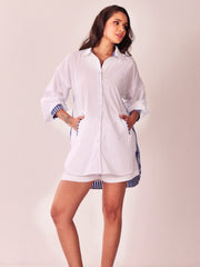 A woman modeling a white button-up shirt dress with blue and white striped trim on the cuffs and side hem, paired with matching striped shorts, against a plain light beige studio background.