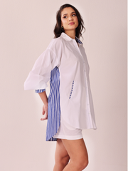 A woman modeling a white oversized button-up shirt dress with blue and white striped side panels and striped inner collar, photographed against a plain light beige studio background.