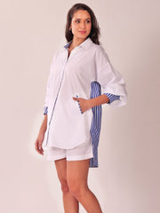A woman modeling a white oversized shirt with blue and white striped accents on the inner collar, cuffs, and side panel, paired with matching white shorts, against a plain light beige studio background.