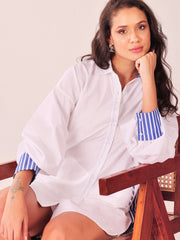 A woman wearing a white button-up shirt with blue and white striped cuffs, sitting on a wooden chair against a plain light background.