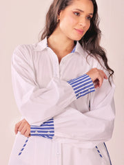 A woman wearing a white long-sleeve button-up shirt with blue and white striped cuffs and hem detail, posed against a light beige background.