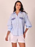 A woman modeling a light blue button-up shirt with navy blue striped chest pocket details and white shorts, set against a plain beige studio background.
