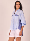 A woman modeling a light blue button-up shirt with blue and white striped details on the chest and sides, paired with white shorts, against a solid beige background.