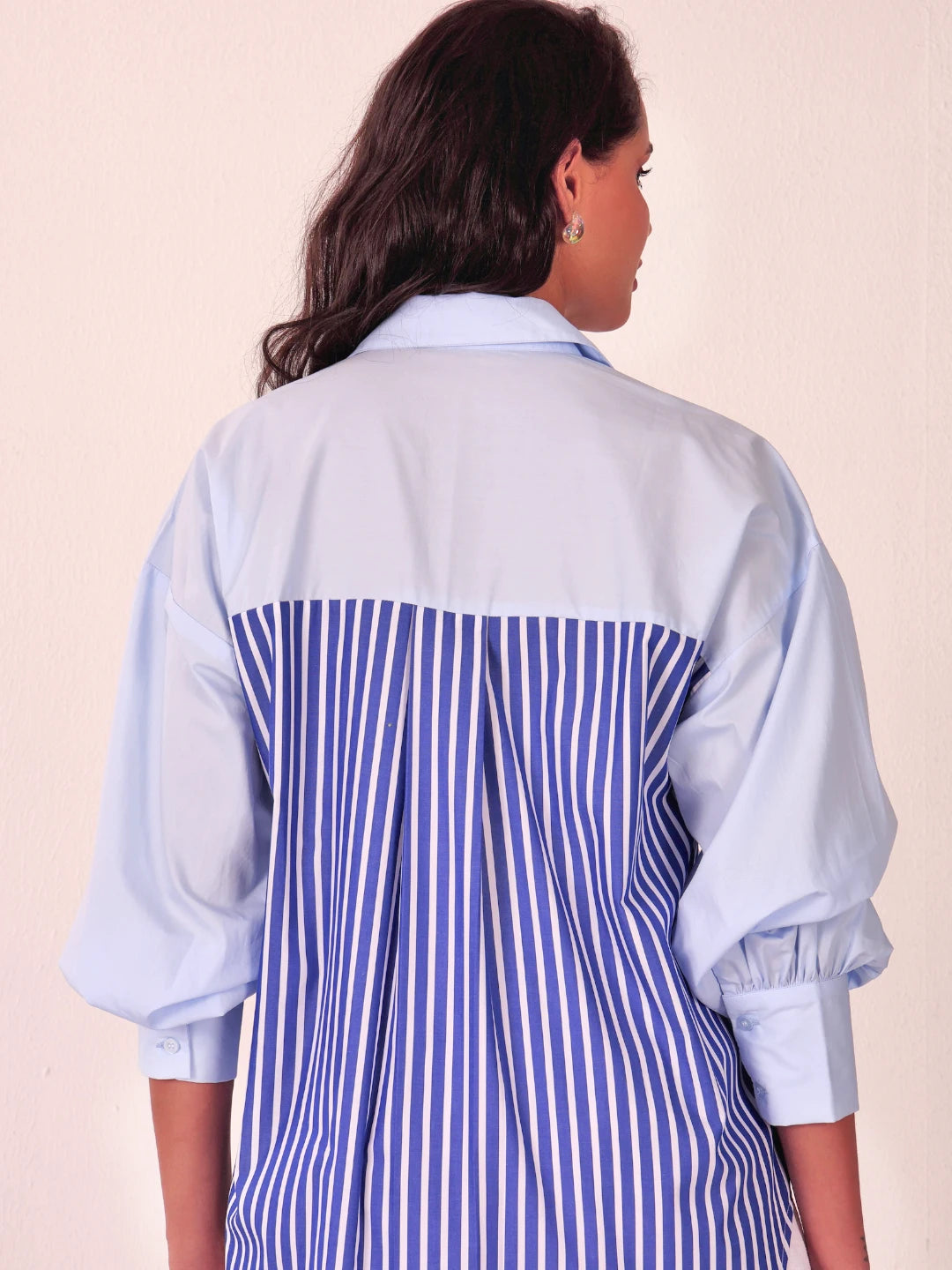 A woman shown from the back wearing a light blue collared shirt with a blue and white vertically striped panel across the back and gathered elbow-length sleeves.