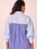 A woman shown from the back wearing a light blue collared shirt with a blue and white vertically striped panel across the back and gathered elbow-length sleeves.