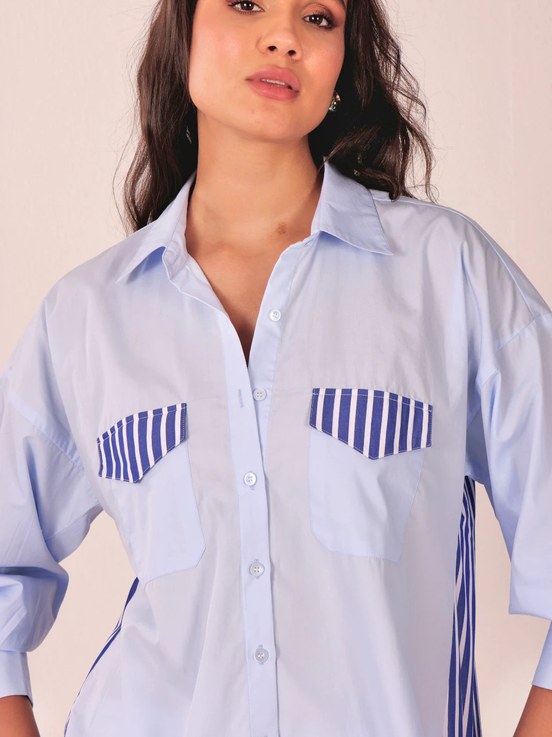 A woman wearing a light blue button-up shirt with blue and white striped chest pocket flaps and side details, photographed against a plain light beige background.