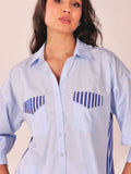 A woman wearing a light blue button-up shirt with blue and white striped chest pocket flaps and side details, photographed against a plain light beige background.