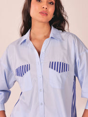 A woman wearing a light blue button-up shirt with blue and white striped chest pocket flaps and side details, photographed against a plain light beige background.