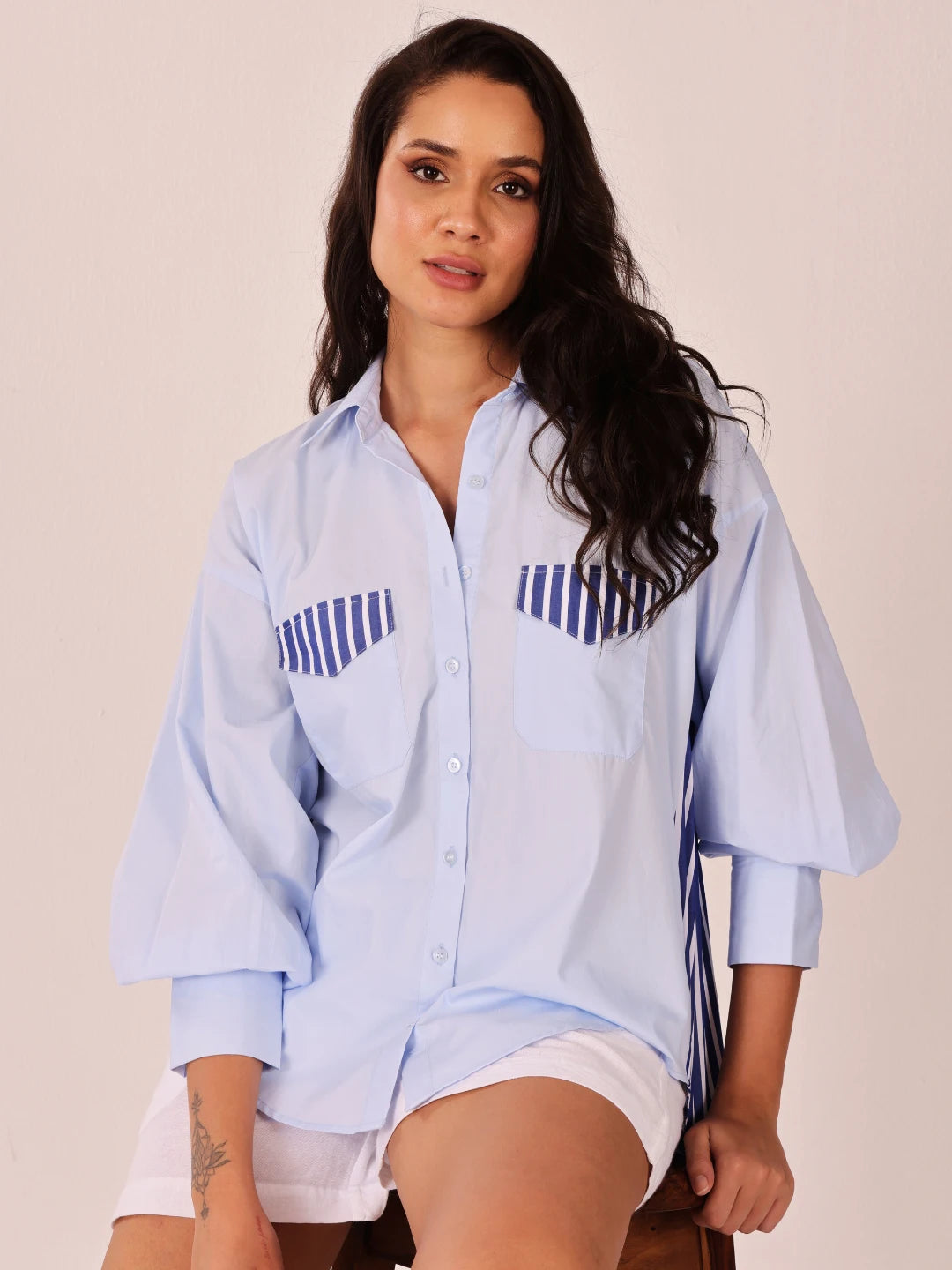 A woman wearing a light blue button-up shirt with navy blue striped chest pocket details, paired with white shorts, posed against a solid light beige background.