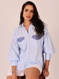 A woman wearing a light blue button-up shirt with navy blue striped chest pocket details, paired with white shorts, posed against a solid light beige background.