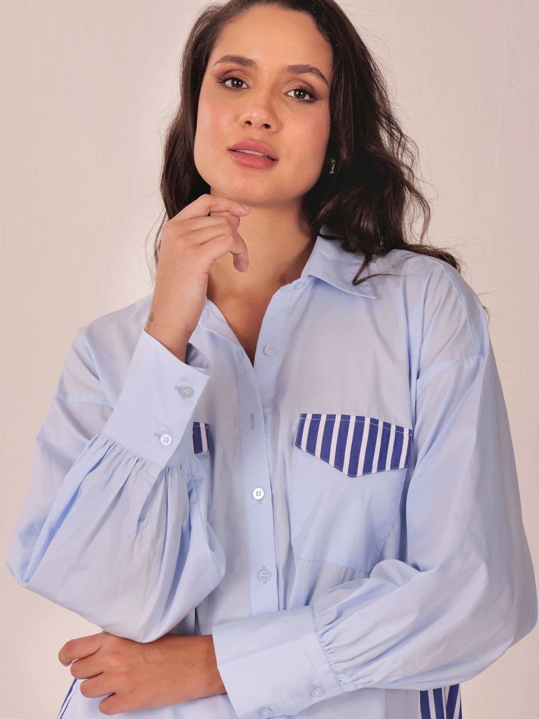 A woman wearing a light blue long-sleeve button-up shirt with a navy blue and white striped chest pocket, posed against a plain beige background.
