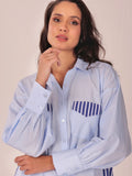 A woman wearing a light blue long-sleeve button-up shirt with a navy blue and white striped chest pocket, posed against a plain beige background.