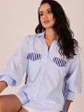 A woman wearing a light blue button-up shirt with navy blue and white striped accents on the chest pockets and side seams, paired with white shorts, set against a plain beige background.