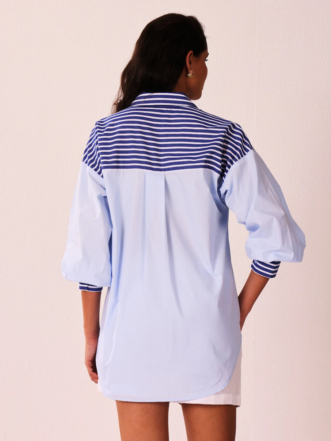 A woman viewed from the back wearing a light blue oversized shirt dress with a navy and white striped yoke and striped cuffs, against a pale pink background.