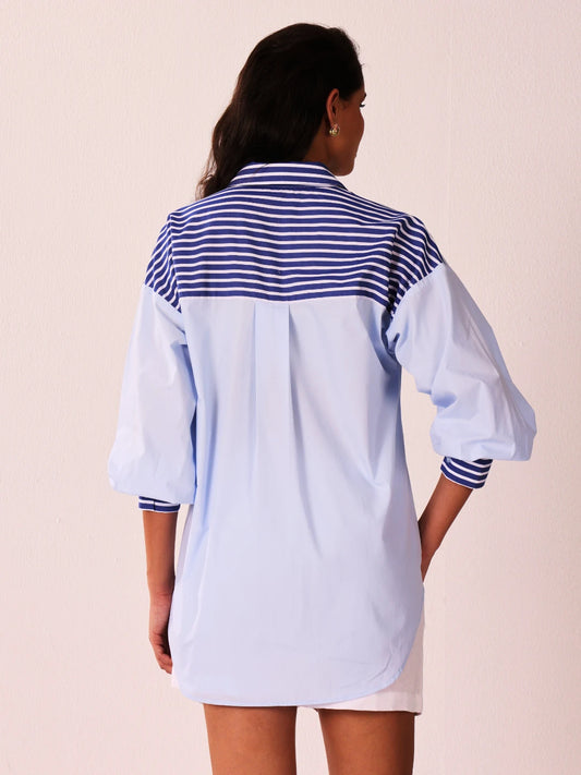 A woman viewed from the back wearing a light blue oversized shirt dress with a navy and white striped yoke and striped cuffs, against a pale pink background.