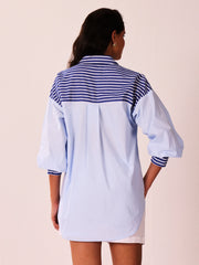 A woman viewed from the back wearing a light blue oversized shirt dress with a navy and white striped yoke and striped cuffs, against a pale pink background.