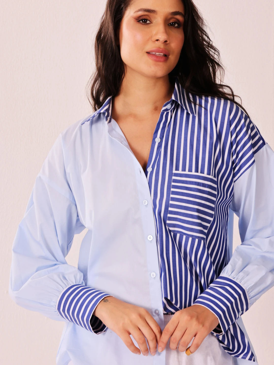 A woman wearing a long-sleeve button-up shirt with a split design: one half is solid light blue and the other half features vertical blue and white stripes. The image is cropped from the chest up against a plain light beige background.