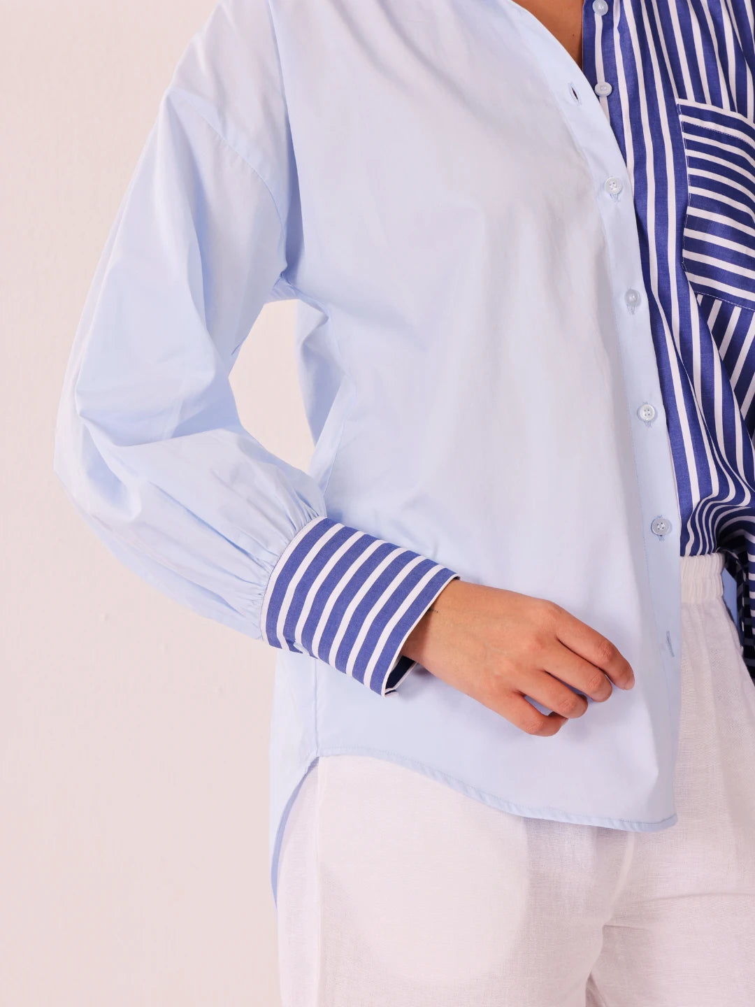 A close-up of a person wearing a light blue button-up shirt with blue and white striped cuffs, layered over a matching blue and white striped top and white pants.
