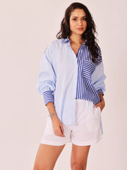 A woman modeling a light blue button-up shirt with blue and white vertical striped panels on the front and sleeves, paired with white shorts, against a solid light beige background.