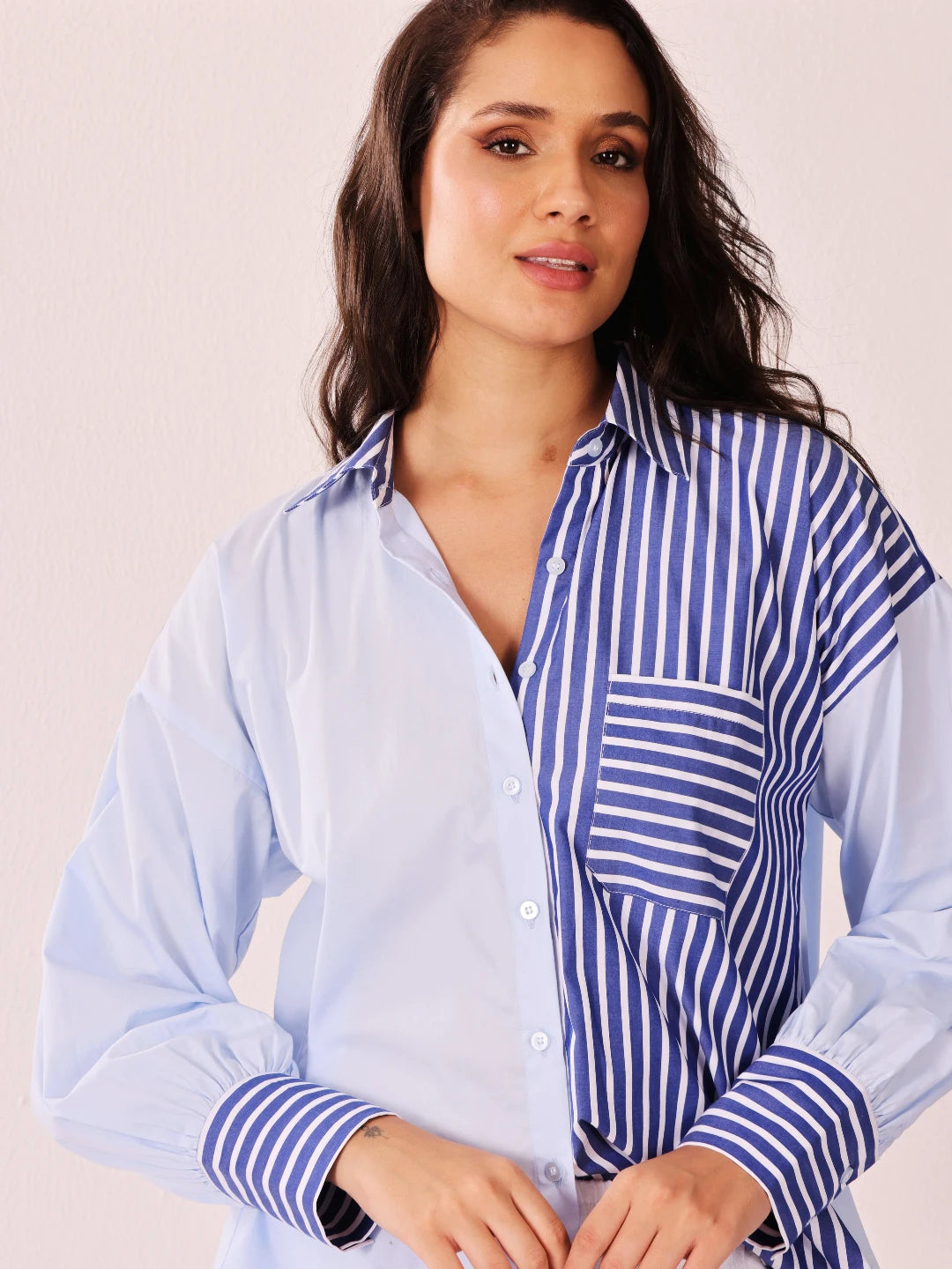 A woman wearing a long-sleeve button-up shirt with a split design: one half is solid light blue, and the other half features blue and white vertical stripes. The shirt has a pointed collar and a chest pocket.