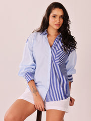 A woman wearing a light blue button-up shirt with blue and white vertical stripes on the front panel and pocket, paired with white shorts, posed against a solid light beige background.