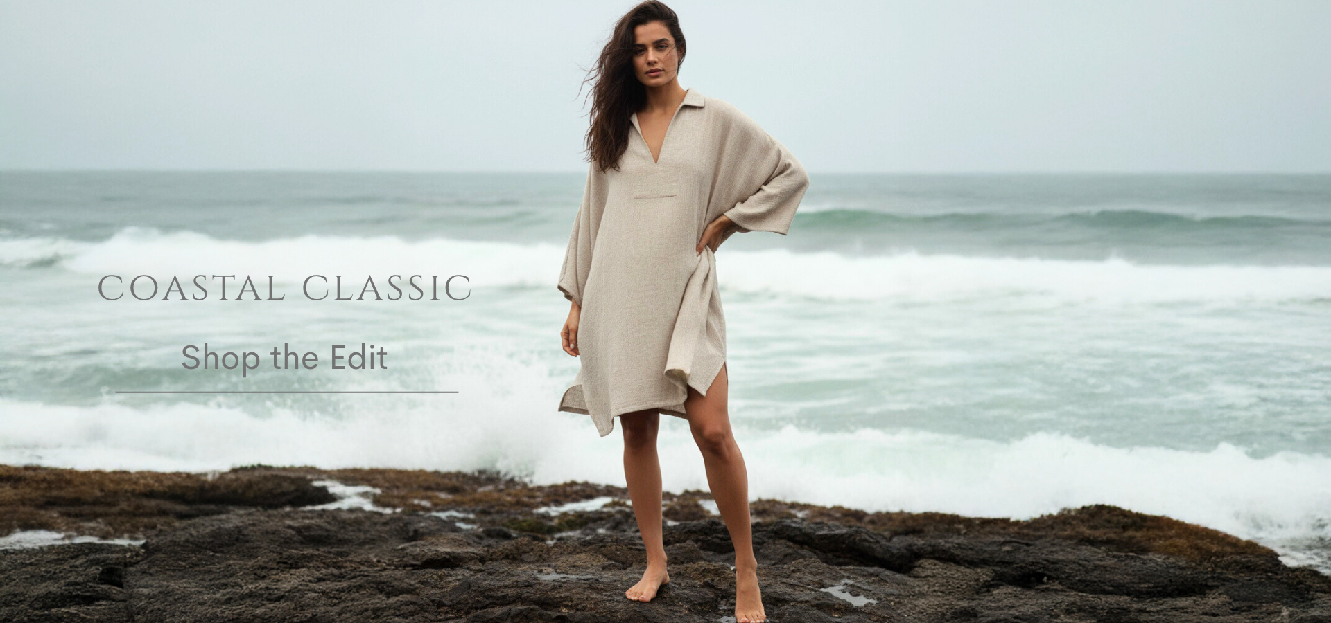 Woman in a beige dress standing on a rocky beach with ocean waves in the background