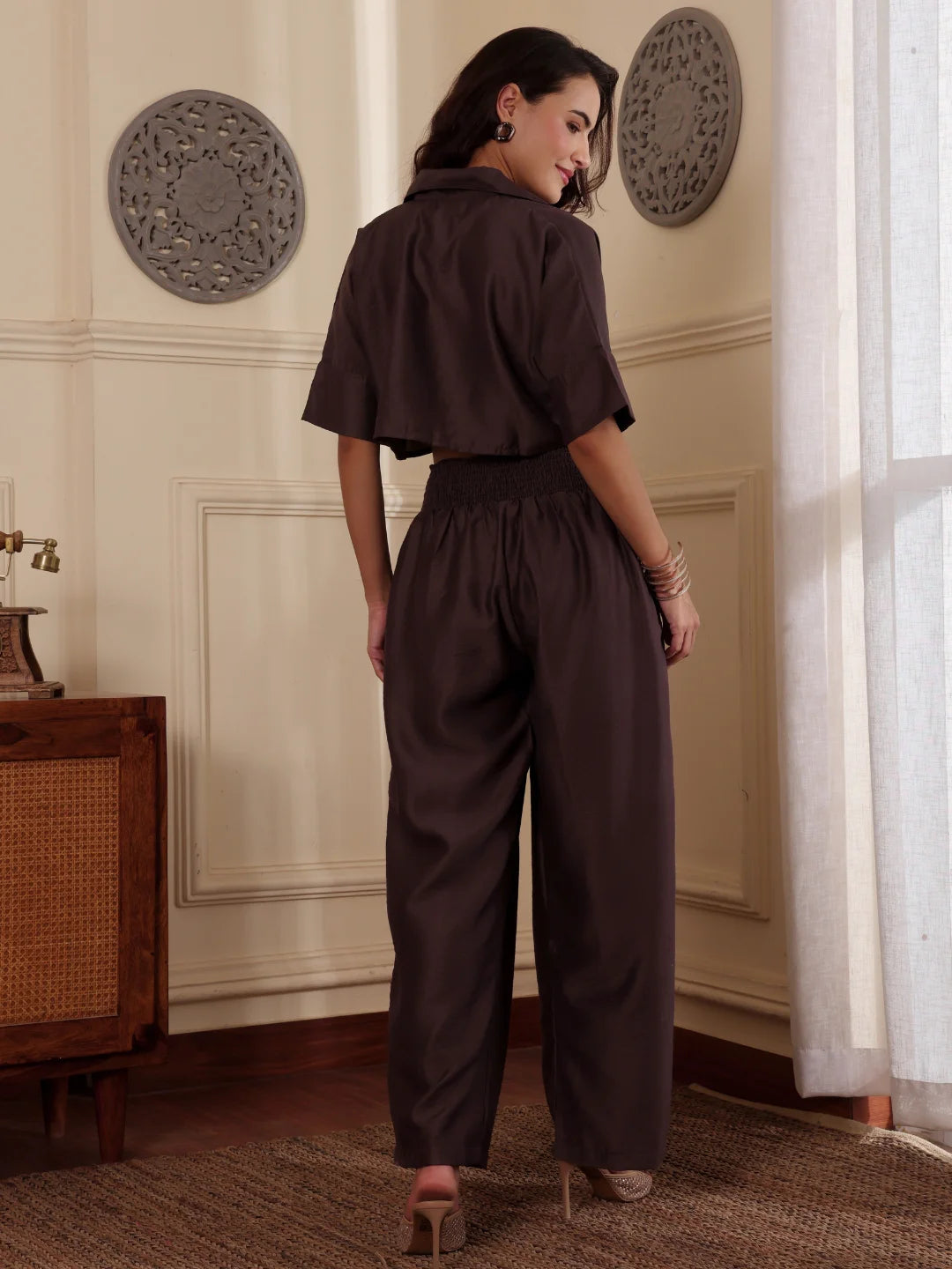 Back shot of a Woman wearing a brown co-ord set with crop shirt and flared Pant outfit standing in a room with a neutral wall and window.