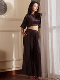 Woman wearing a brown co-ord set with crop shirt and flared Pant outfit standing in a room with a neutral wall and window.