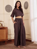 Woman wearing  a brown co-ord set with crop shirt and flared Pant outfit standing in a room with a neutral wall and window.
