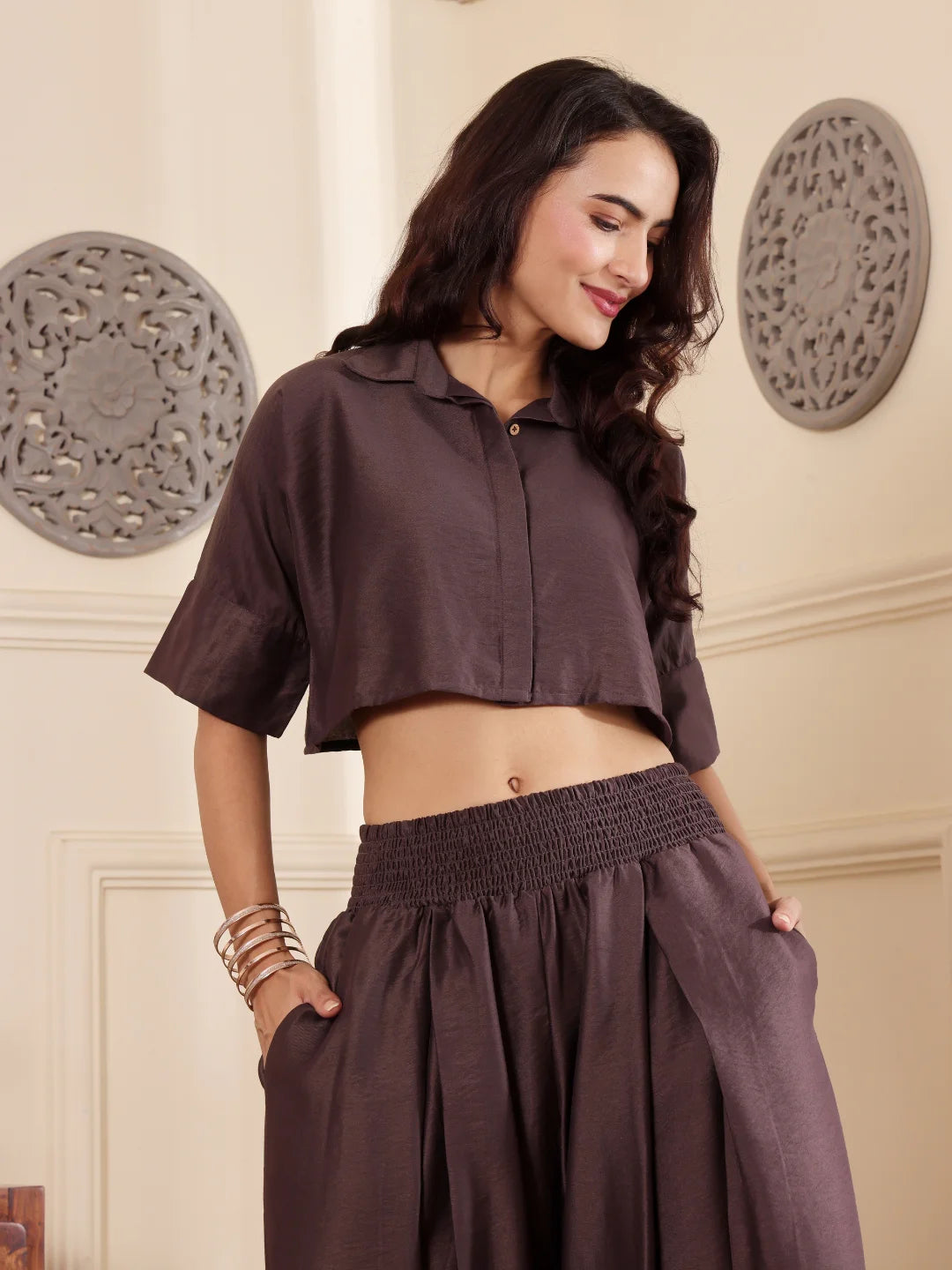 Woman wearing a brown co-ord set with crop shirt and flared Pant outfit , with both hand in side pockets, standing in a room with a neutral wall and window.