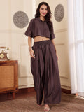 Woman walking wearing a brown co-ord set with crop shirt and flared Pant outfit standing in a room with a neutral wall and window.
